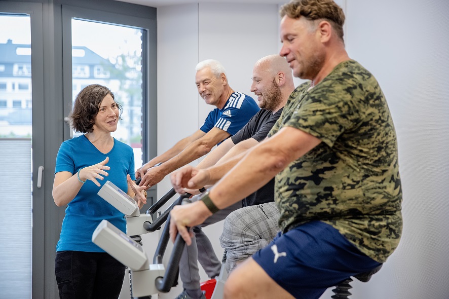 Mehrere Patienten auf einem Ergometer werden von einer Physiotherapeutin betreut. Mehrere Patienten auf einem Ergometer werden von einer Physiotherapeutin betreut.